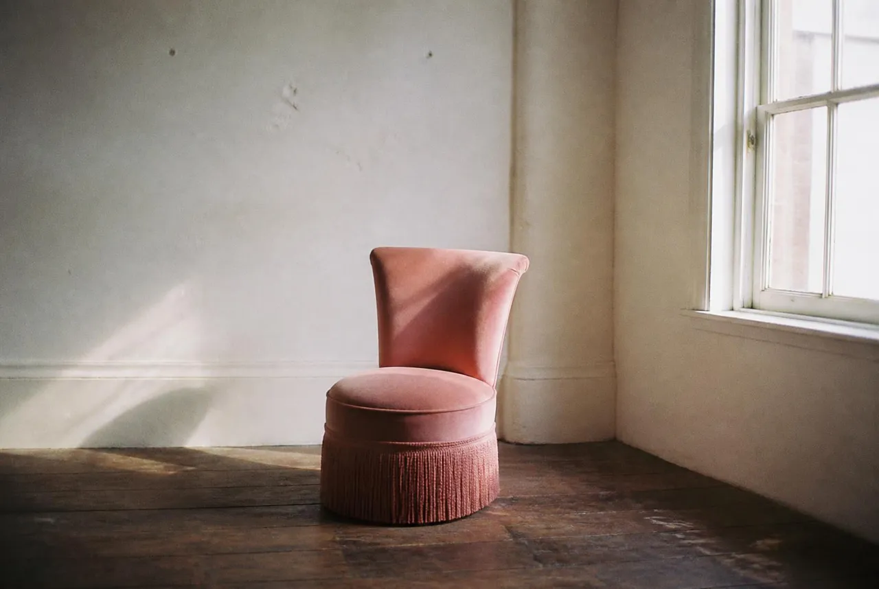 A single pink chair alone in an empty white studio room, lit by one window